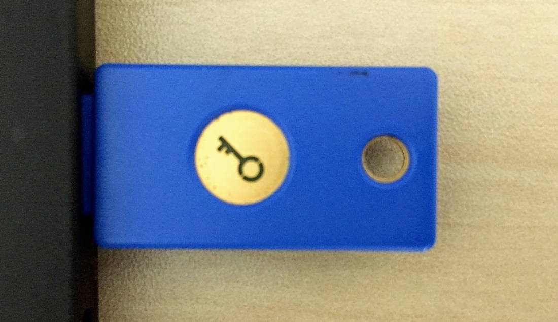 Photo of a Yubi FIDO U2F key in a black laptop. The Key is blue with a gold key emblem on a wood grain backgrond